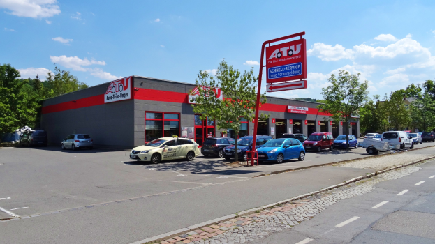 Autohaus mit parkenden Autos vorne, umgeben von Gebäuden, Bäumen, Strommasten, Schildern und einer Straße unter einem bewölkten Himmel.