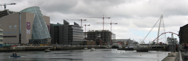 Ein Fluss mit Schiffen, eine Brücke, Gebäude, Baukräne und ein Himmel im Hintergrund.