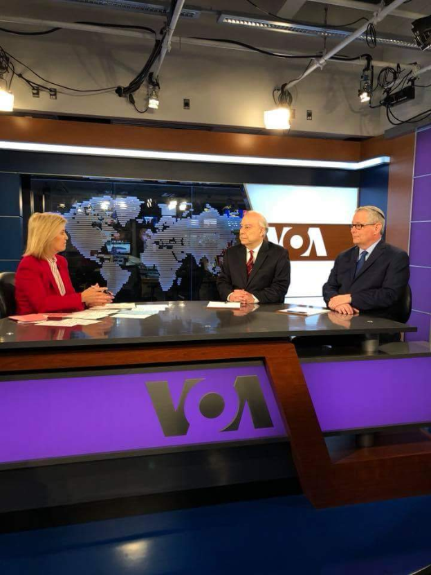 Drei Personen, zwei Männer in Blazern und Krawatten und eine Frau in einem roten Kleid sitzen an einem Schreibtisch mit Papieren und Gegenständen, diskutieren vor einem Fernseher mit einem Bildschirm und Lichtern im Hintergrund.