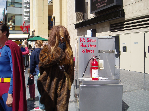 Eine Menge Menschen, einige in Kostümen, versammelte sich vor Gebäuden mit sichtbarer Beschriftung, einer Box mit Schrift und einem Feuerlöscher.