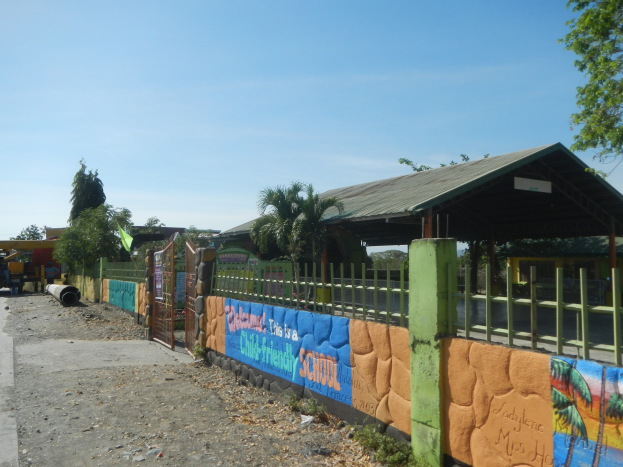 Schulgebäude mit einer Wandmalerei, umgeben von einem Zaun, einem Schuppen, Bäumen, Fahrzeugen, Pfählen und einem bewölkten Himmel.