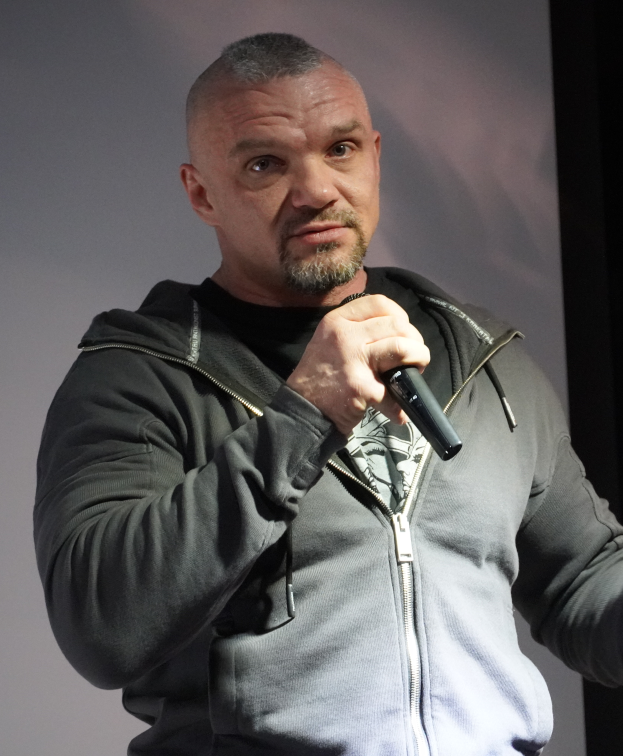 Ein Mann in einer schwarzen Kapuzenjacke, schwarzem Anzug und weißem Hemd hält ein Mikrofon und steht selbstbewusst vor einer Wand.