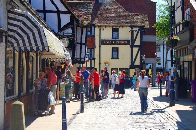 Eine Straße mit Pfählen und Gebäuden auf beiden Seiten, Menschen auf der linken Seite mit einem Handwagen, ein Mann und eine Frau auf der rechten Seite in der Nähe eines lilafarbenen Müllcontainers, mit Gebäuden, Bäumen und Blumentöpfen im Hintergrund.