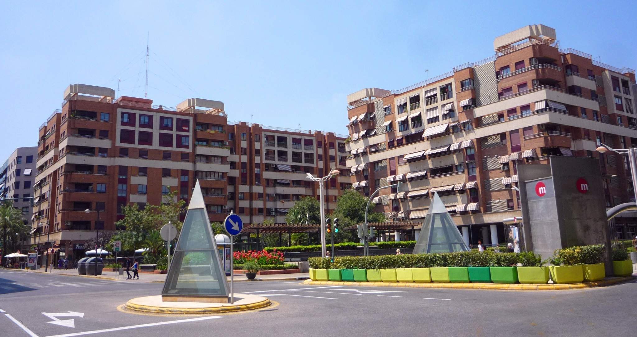 Eine Stadtstraße mit einem Denkmal im Vordergrund, hohen Gebäuden im Hintergrund, Straßenmöblierung, Grünflächen, Fußgängern und einem klaren blauen Himmel.