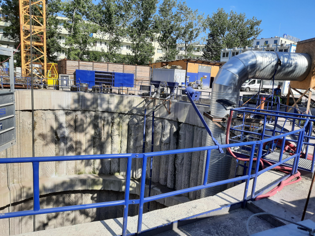 Eine Baustelle an einer Kläranlage mit einem großen Metallrohr in der Mitte, umgeben von Geländern, Rohren und Maschinen, mit Gebäuden, Bäumen und einem Turm im Hintergrund.