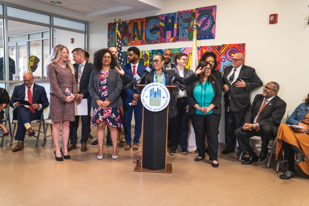 Eine Gruppe von Menschen steht und sitzt in einem Raum mit einem Podium, Mikrofon und einer Tafel mit der Aufschrift "Sancho-Stadtrat tritt dem Stadtrat bei", mit Flaggen, Feuerlöscher und Deckenleuchten an der Rückwand.