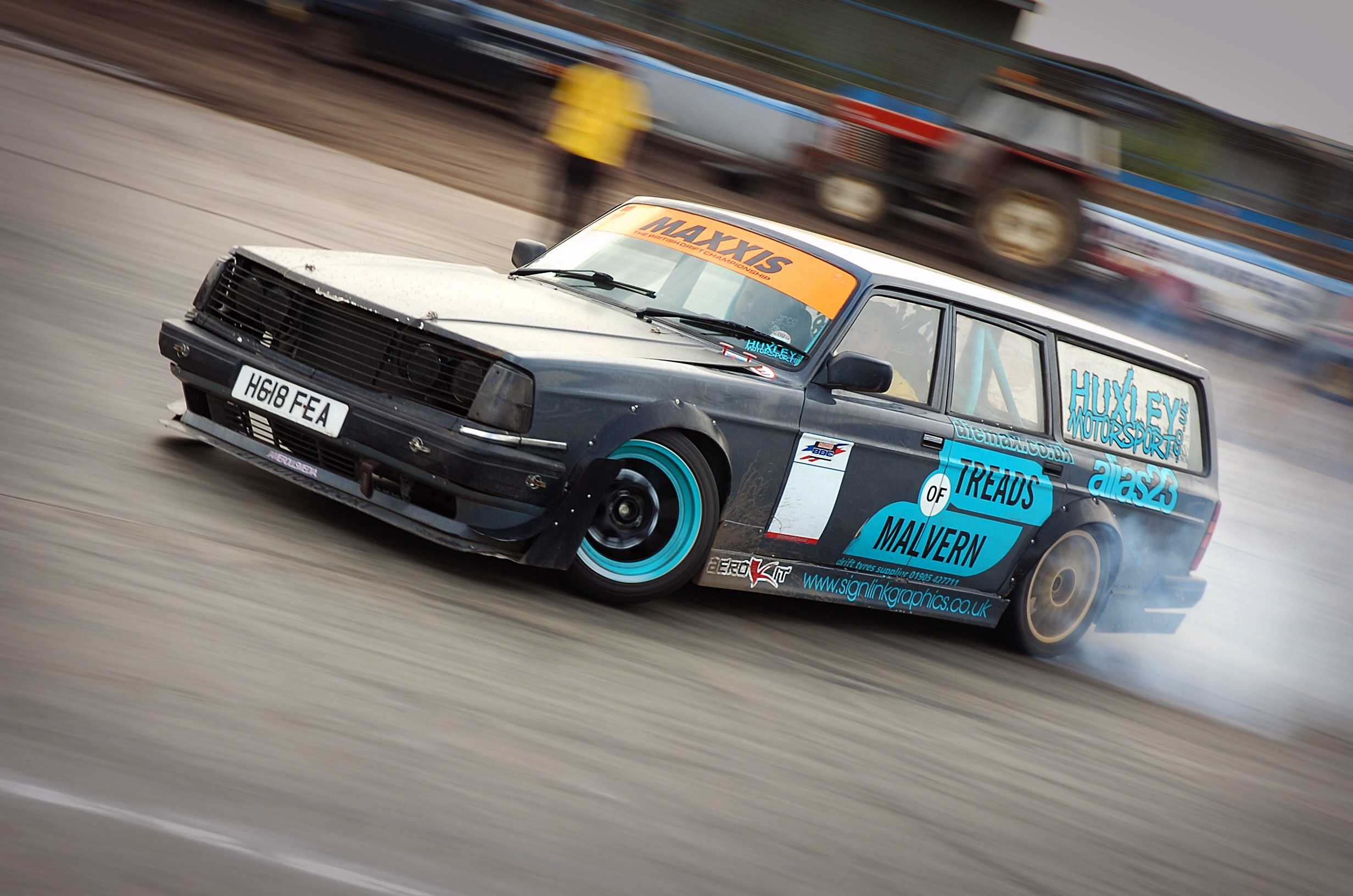 Ein Rennwagen, der Rauch hinter sich herzieht, während er auf einer Rennstrecke umgeben von anderen Fahrzeugen und Zuschauern fährt.