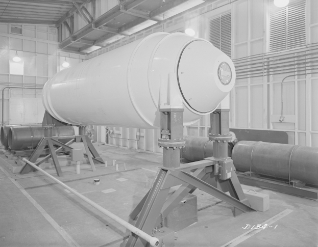 Schwarzes und weißes Foto einer großen zylindrischen Wasserstoffbombe auf einem Ständer in einer Fabrik mit Rohren, Lampen und einer Wand im Hintergrund.