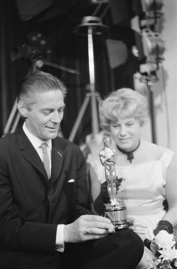 Ein Mann in einem schwarzen Blazer, weißem Hemd und Krawatte hält eine Oscar-Statue in der Hand und sitzt neben einer Frau, die einen Blumenstrauß hält, beide in formeller Kleidung, mit Scheinwerfern im Hintergrund, in Schwarz-Weiß.