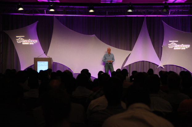 Ein Mann in einem blauen Hemd steht auf einer Bühne mit einem schwarzen und weißen Vorhang und einem Metallrahmen mit einer Spotlight darüber und spricht zu einem sitzenden Publikum.