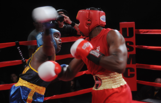 Zwei Boxer kämpfen im Ring, während ein Fotograf und Zuschauer im Hintergrund unter dramatischer Beleuchtung zu sehen sind.