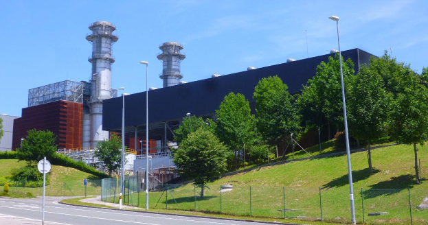 Großes Gebäude mit zahlreichen Fenstern, als Kraftwerk durch Beschilderung identifiziert, umgeben von Straßeninfrastruktur, Fahrzeugen, Grünflächen und einem bewölkten Himmel.