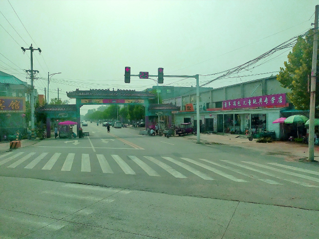 Stadtstraßenkreuzung mit Zebrastreifen, Ampel, Fahrzeugen, Strommasten, Gebäuden, Beschilderung, Bäumen, Schirmen und einem Bogen im Hintergrund unter einem sichtbaren Himmel.