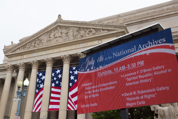 Das Nationalarchiv-Gebäude in Washington, DC, mit einer Tafel mit Text, Flaggen, Laternenpfählen, Bäumen, Skulpturen und einem klaren blauen Himmel.