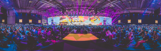 Eine große Menschenmenge sitzt auf Stühlen, die einer hell erleuchteten Bühne zugewandt sind, mit mehreren stehenden Individuen, wahrscheinlich auf einem Online-Marketing-Rockstars-Event, umgeben von Bildschirmen.