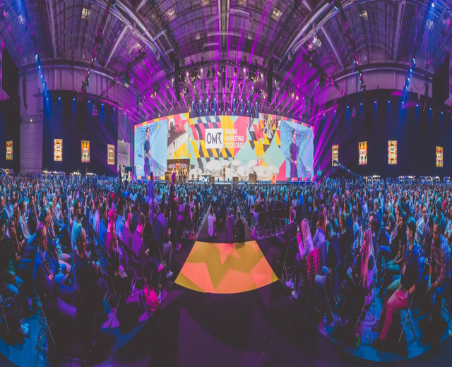 Eine große Menschenmenge sitzt auf Stühlen, die einer hell erleuchteten Bühne zugewandt sind, mit mehreren stehenden Individuen, wahrscheinlich auf einem Online-Marketing-Rockstars-Event, umgeben von Bildschirmen.