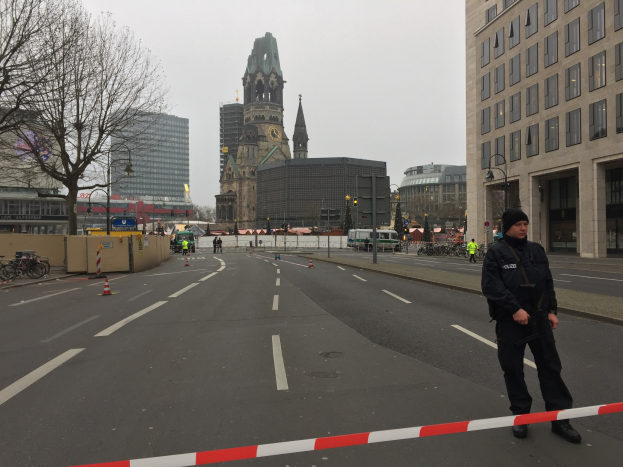 Polizeibeamter in schwarzer Uniform und Mütze auf einer Straße in Berlin mit Verkehrskegeln, Fahrrädern, Laternenmasten, Bäumen, Gebäuden, einem Kirchturm und einem klaren blauen Himmel im Hintergrund.