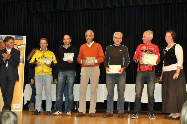 Eine Gruppe von Menschen auf einer Bühne mit Urkunden in der Hand, im Hintergrund eine Wand und ein Tisch sowie ein schwarzer Vorhang.