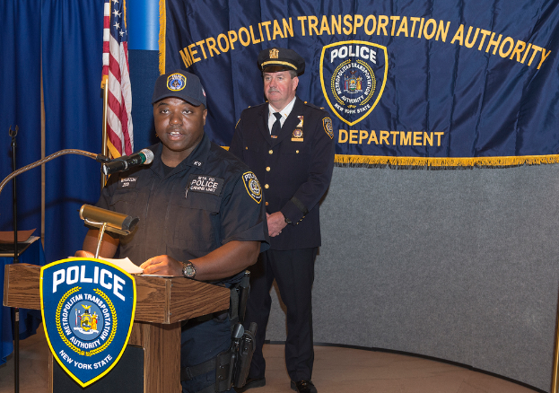 Zwei Polizeibeamte stehen vor einem Rednerpult, einer h├Ąlt Papiere und spricht in ein Mikrofon, mit einer Fahne, einem Banner und einem Tisch in der N├Ąhe und einer Tafel mit Symbolen und Text im Hintergrund.
