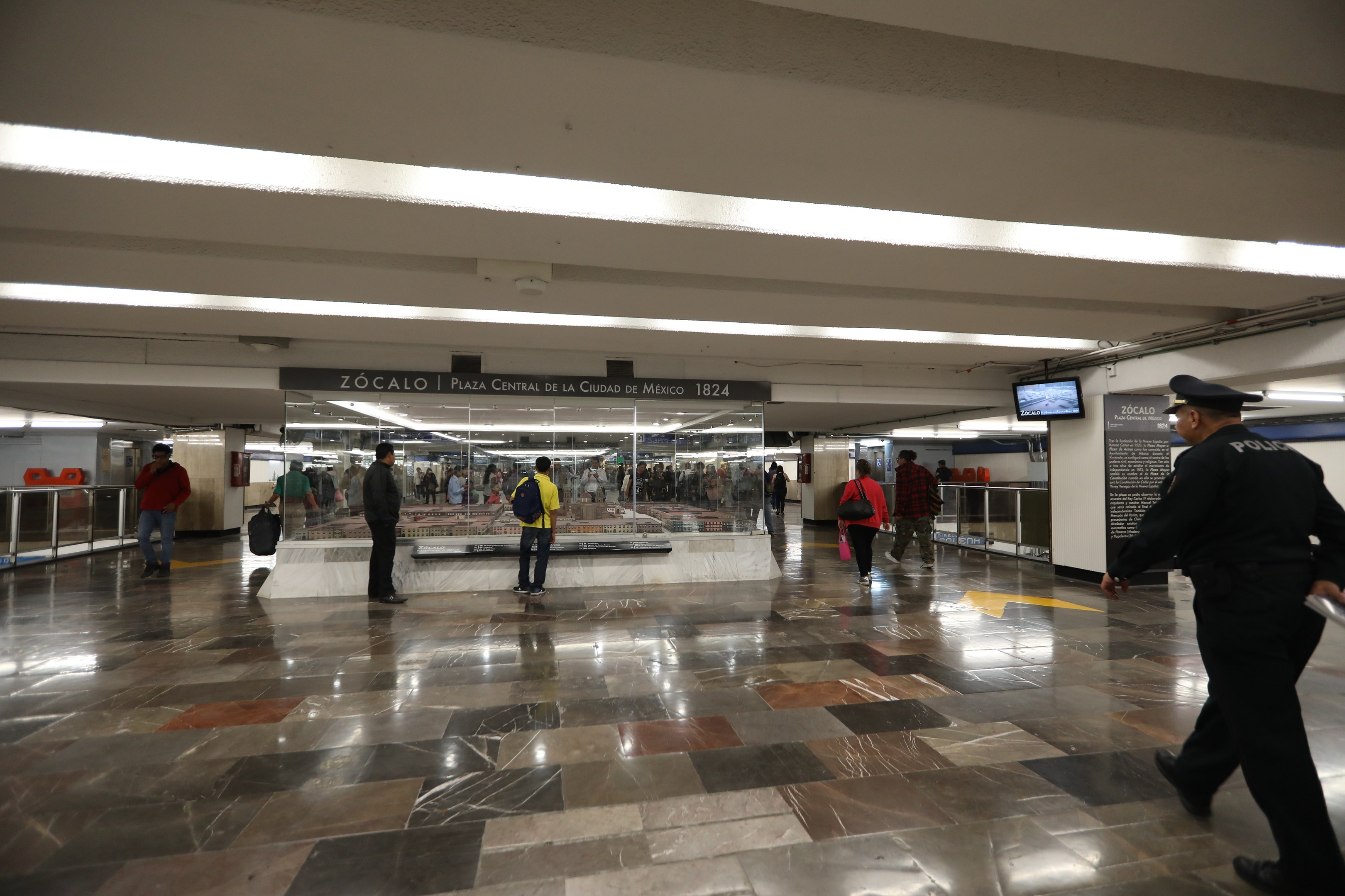 Eine Gruppe von Menschen geht durch eine belebte U-Bahn-Station mit einem Polizeibeamten im Vordergrund, über den Bahnsteigen sind Texte und Bildschirme zu sehen, sowie verschiedene bauliche Elemente wie Geländer und Lampen.