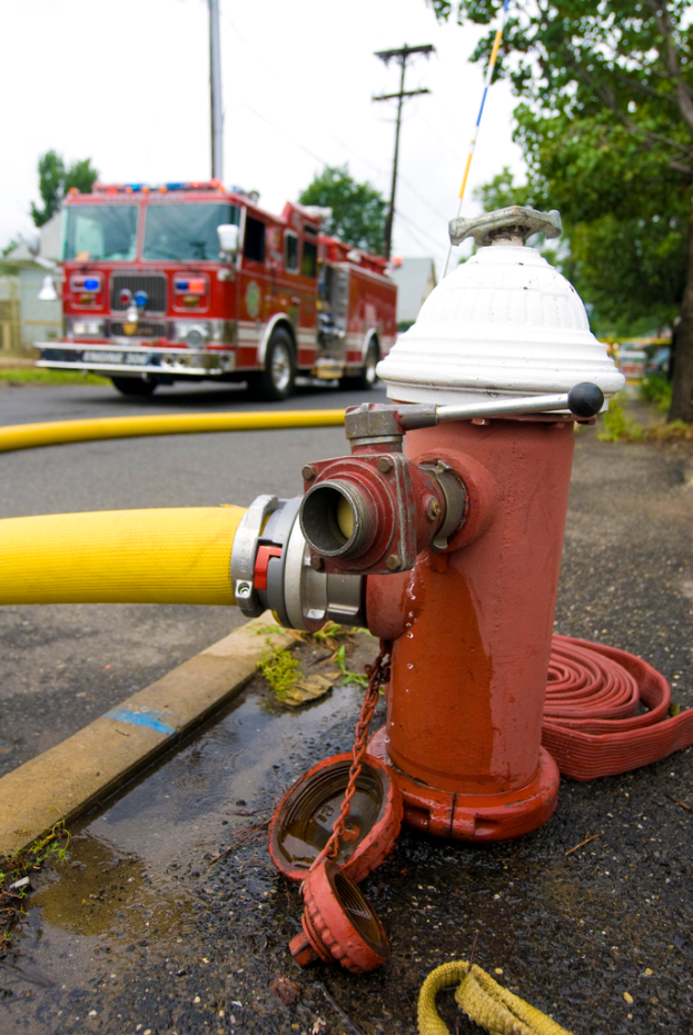 Ein Feuerhydrant und ein gelber Schlauch im Vordergrund mit einem Bus, Stromkabel, Masten und Bäumen im Hintergrund.