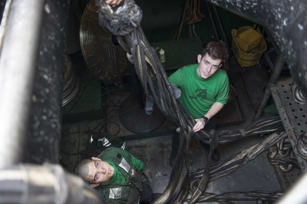 Zwei Männer in grünen Hemden reparieren eine Maschine umgeben von Werkzeugen, Drähten, einem Ventilator und einer Flasche.