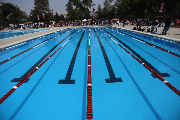 Großes Schwimmbad mit Bahntrennern, umgeben von Menschen mit Taschen und Gegenständen, mit Zelten, Bannern, Pfosten, einem Schild und einem bewölkten Himmel im Hintergrund.