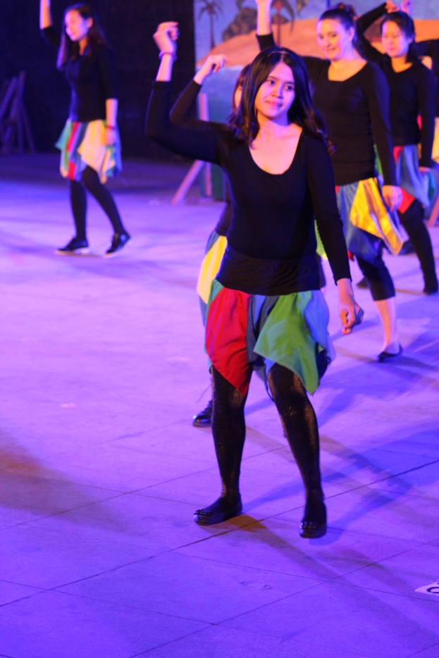 Eine Gruppe junger Frauen tanzt auf einer Bühne mit einem Gemälde im Hintergrund, alle tragen Schuhe.