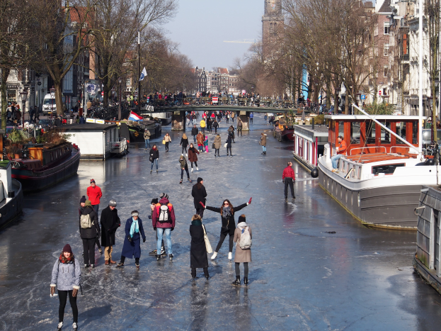 Eine Gruppe von Menschen, die auf einem zugefrorenen Kanal in Amsterdam Eislaufen, umgeben von Booten, Bäumen, Gebäuden, Laternenmasten, Flaggen und einer Brücke unter einem klaren Himmel.