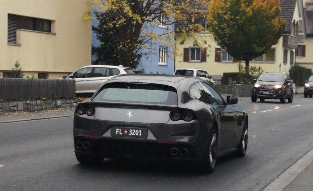 Roter Ferrari GTC4Lusso fährt auf einer Straße mit Gebäuden, Bäumen und einem Zaun im Hintergrund unter einem sichtbaren Himmel.