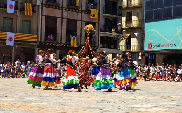 Eine Gruppe von Frauen in bunten Kleidern tanzt auf der Straße, umgeben von einer Menschenmenge, mit Gebäuden, Gelöndern, Balkonen, Flaggen, einem Laternenmast und einer Tafel mit Aufschrift im Hintergrund.