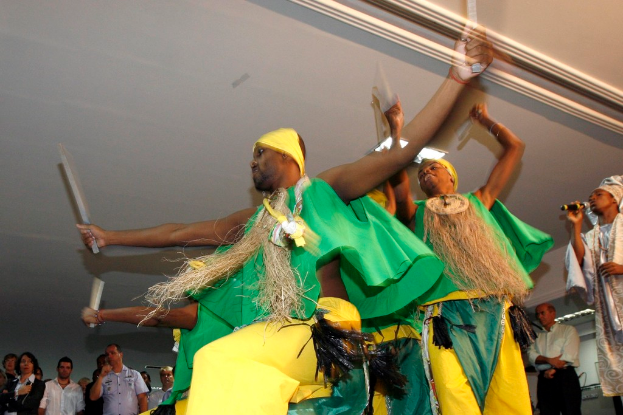 Männer im Vordergrund führen einen Tribaltanz auf, rechts steht ein Mann mit Mikrofon, im Hintergrund stehen einige Personen unter einer Decke.