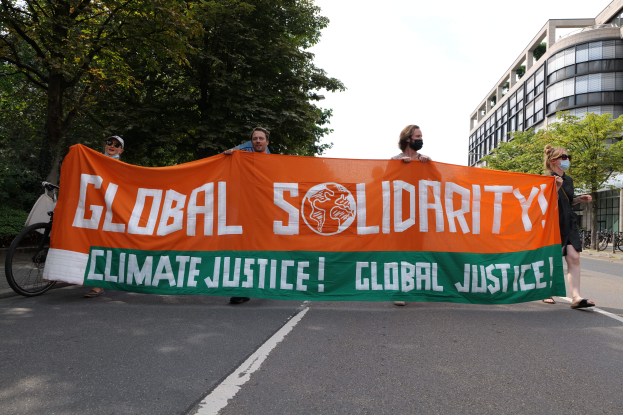 Eine Gruppe von Menschen marschiert eine baumbestandene Straße entlang und hält ein Transparent mit der Aufschrift "Globale Solidarität: Klimagerechtigkeit". Fahrräder sind entlang der Straße geparkt und im Hintergrund sind Gebäude zu sehen.