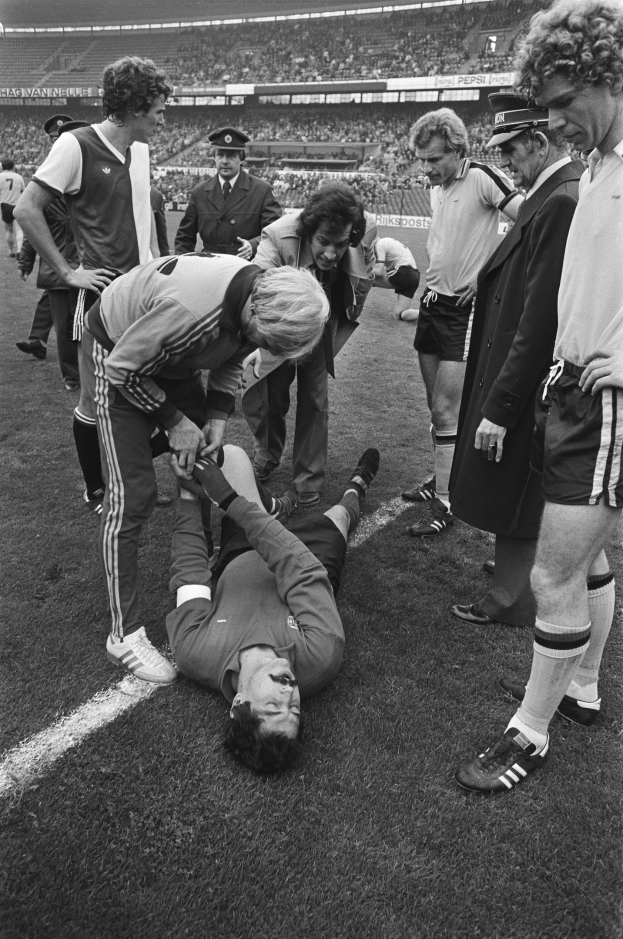 Schwarzes und wei├čes Bild eines Fussballspielers, der auf dem Boden liegt und von einer Gruppe M├Ąnner umgeben ist, mit einer Menge im Hintergrund eines Stadions.