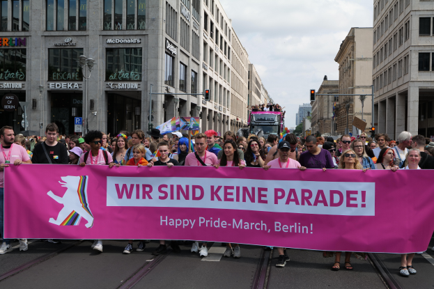 Eine Gruppe von Menschen geht auf einer Straße in Berlin, Deutschland, mit einer pinken Fahne, auf der 'Happy Pride March' steht, vorbei, während Gebäude, Laternenpfähle und Verkehrszeichen die Straße säumen und ein bewölkter Himmel im Hintergrund zu sehen ist.