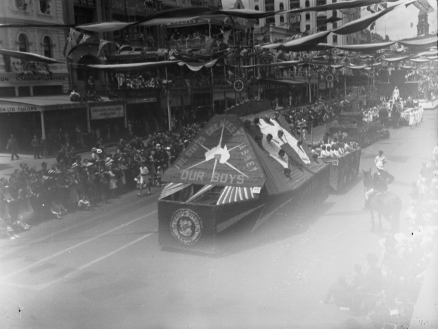 Ein Schwarz-Weiß-Foto eines Umzugs mit einem Wagen, Menschen auf der Straße, einige zu Pferd, Gebäude mit Fenstern im Hintergrund und sichtbare Fahnen.