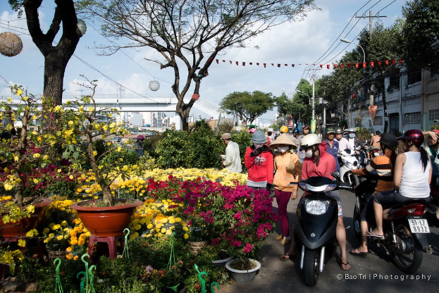 Gruppe von Menschen mit Helmen und Muetzen, einige gehen und andere fahren mit Faehrraedern auf einer Strasse mit Pflanzen, einem Blumentopf auf einem Hocker, Baeumen, einem Pfahl, Drahten, einer Lampe, einer Brucke und Gebaeuden im Hintergrund.