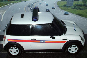 Weißes Polizeiauto auf einer Straße mit mehreren Fahrzeugen, Gras und Bäumen auf beiden Seiten, aufgenommen außerhalb einer Stadt.