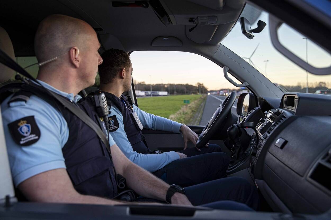 Zwei Polizisten in einem Auto auf einer Straße mit Gras, Bäumen und Pfählen im Hintergrund unter einem klaren blauen Himmel.