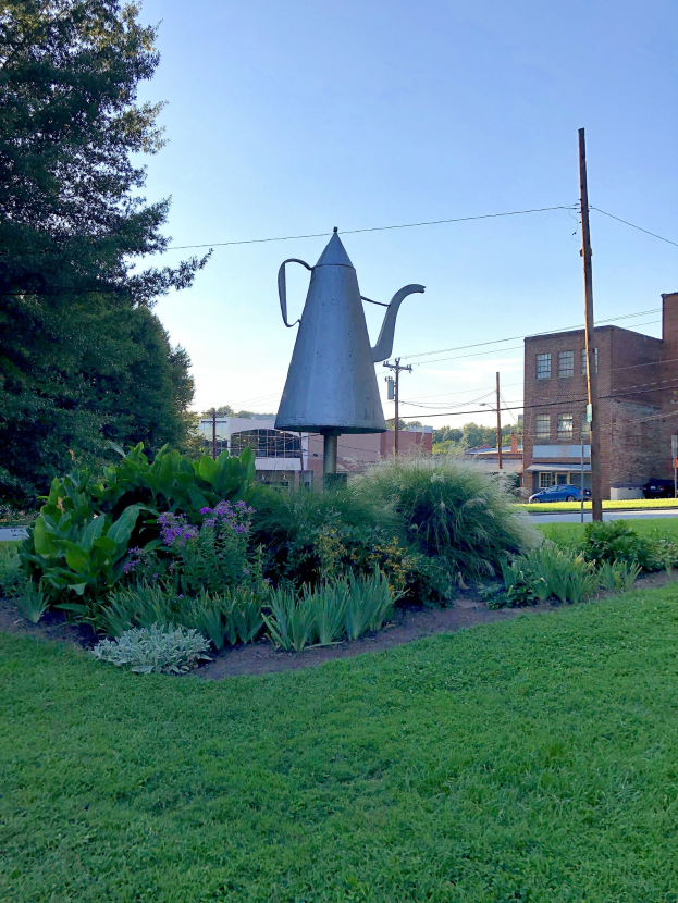 Ein Metallgießer steht auf einem grünen Feld mit Pflanzen, Gras und Bäumen, mit Strommasten, einem Schild, Fahrzeugen, Gebäuden und einem bewölkten Himmel im Hintergrund.