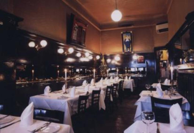 Ein Esstisch mit Gläsern, Kerzen auf Ständern, Servietten, Tellern, Stühlen, wandmontierten Rahmen und Deckenleuchten.