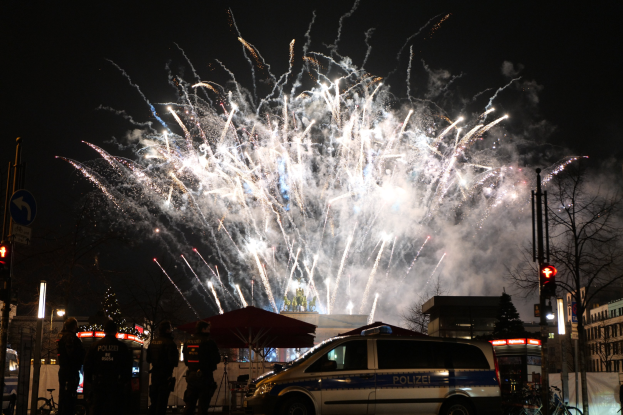 Polizeiauto vor einer Menschenmenge während einer Silvesterfeier in Berlin geparkt, mit farbigen Feuerwerken am Himmel.