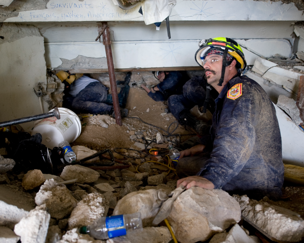 Ein Feuerwehrmann in einem Helm sitzt zwischen Trümmern, darunter Steine und Flaschen, mit einer teilweise sichtbaren Wand im Hintergrund, die wahrscheinlich den Namen des Gebäudes trägt, das von einem israelischen Luftangriff getroffen wurde.