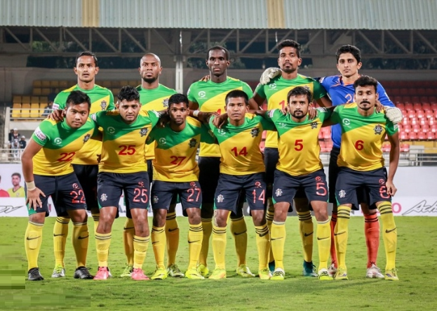 Eine Gruppe von Männern auf einem Fussballfeld, die einen Sieg feiern, umgeben von Werbetafeln, Stühlen, Geländern, Pfählen, Bäumen und einer Hütte im Hintergrund.