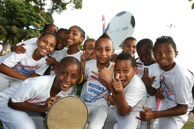 Eine Gruppe strahlender Kinder im Freien mit einem Baum links und dem Himmel im Hintergrund.