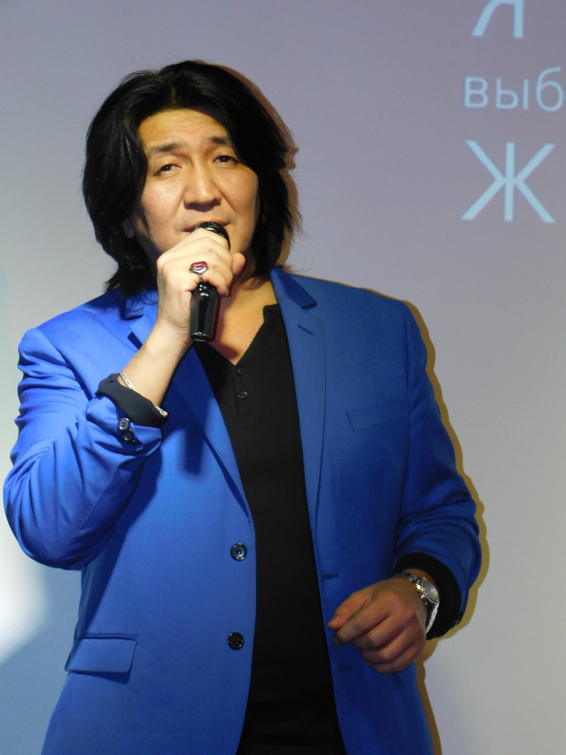 Ein Mann in einem blauen Anzug und einem weißen Hemd, der als Regisseur erscheint, hält ein Mikrofon, während er vor einem Bildschirm mit Text steht und einen ernsten Gesichtsausdruck hat.