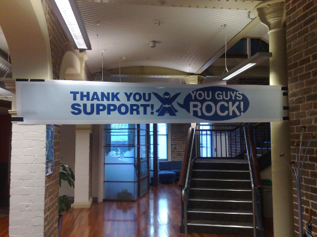 Ein Dankeschön-Banner ist an einer Wand und einer Säule vor einer Treppe angebracht, mit Fenstern im Hintergrund.