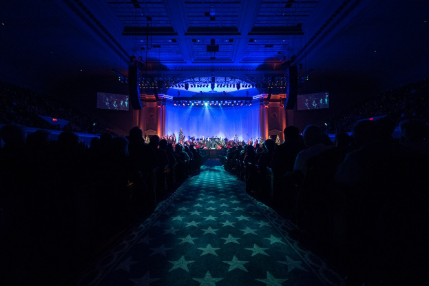 Ein großer Saal mit Menschen, die auf Stühlen sitzen und einer Bühne mit Musikern, ausgestattet mit Deckenleuchten und Fernsehern, typisch für ein Konzerthalle während der Nationalkonvention 2019.