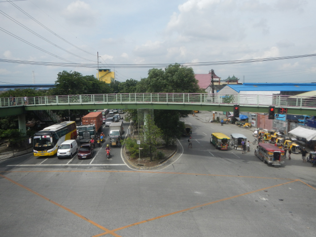 Volles Stadtbild mit Autos, Bussen, Fußgängern, Brücke, Ampeln, Gebäuden, Bäumen, Strommasten und bewölktem Himmel.