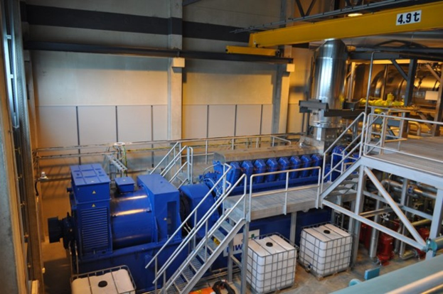 Eine Fabrikinnenszene mit Trägern, einer Treppe mit Geländer, Containern, Lampen, Rohren und Wänden.